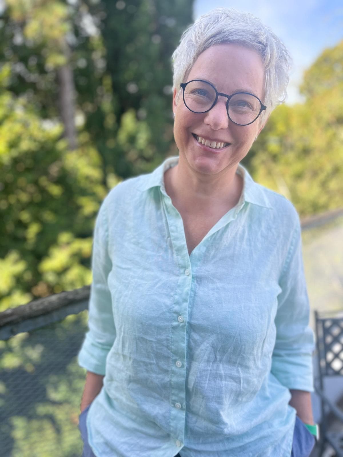 Katharina Hannesschläger in hellblauer Leinenbluse, lächelnd im Freien vor grünen Bäumen