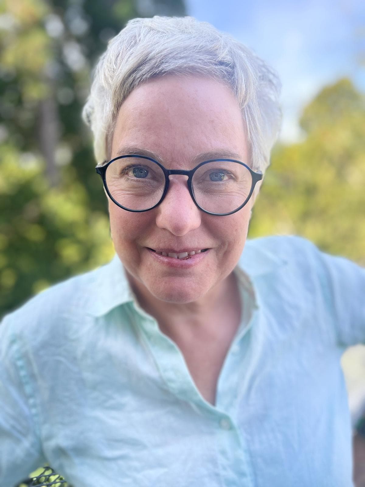 Katharina Hannesschläger in Nahaufnahme, lächelnd mit runder Brille vor sommerlichem Grün